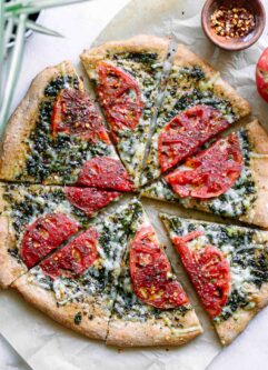 a vegan pizza with pesto sauce and sliced tomatoes on a white table with a bowl of red pepper flakes