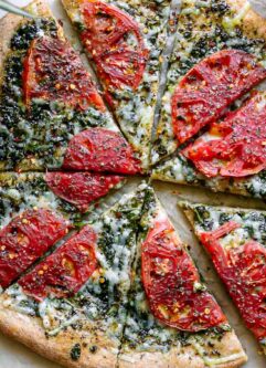 a sliced pesto pizza with roasted tomatoes on a white table with a tomato and a bowl of red pepper