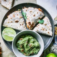 a blue plate with a cut vegan quesadilla with a bowl of guacamole and a lime wedge