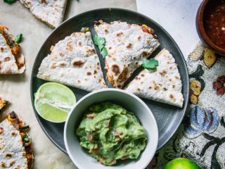 a blue plate with a cut vegan quesadilla with a bowl of guacamole and a lime wedge