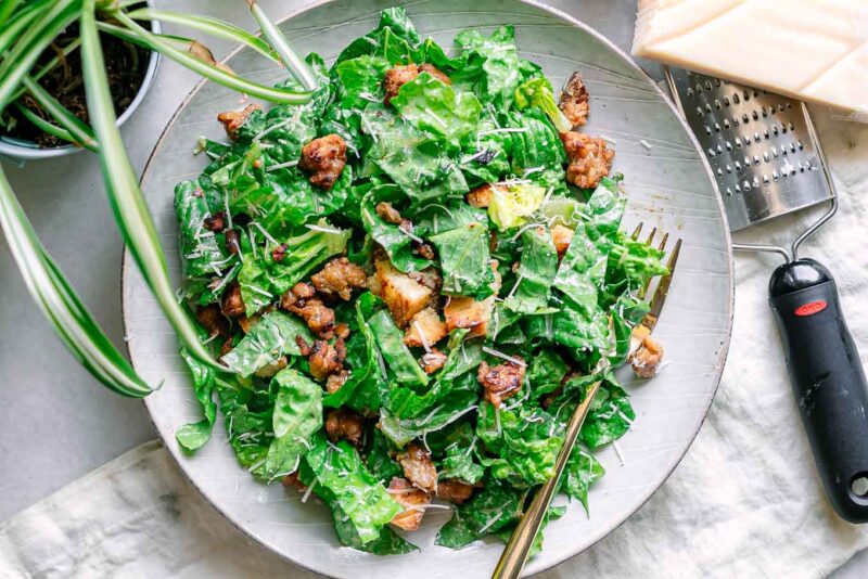 a vegan caesar salad with crumbled crispy tempeh and croutons on a white table with a gold fork