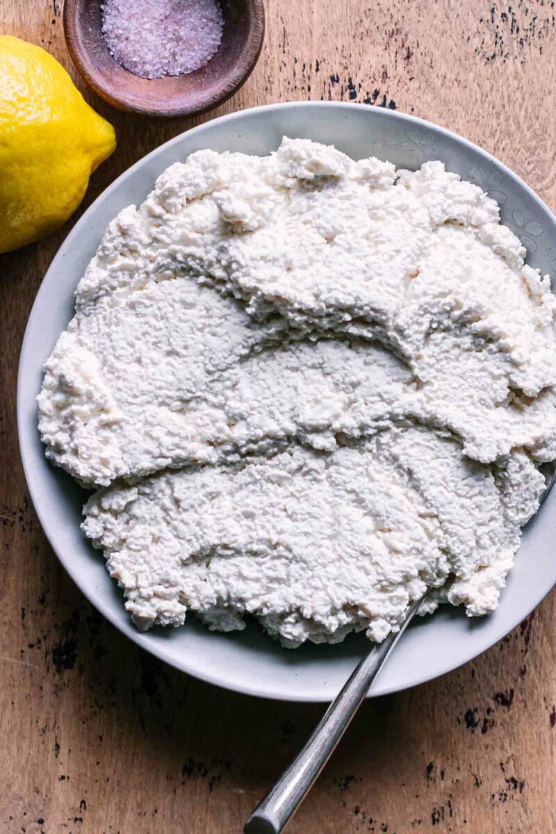 a bowl of tofu ricotta cheese on a wood table with a lemon and a bowl of salt