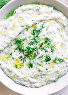 a white bowl with vegan tzatziki sauce on a table with fresh dill and lemons