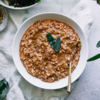 a white bowl with farro and vegetable soup on a white table with sage leaves and a white napkin