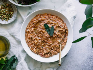 a white bowl with farro and vegetable soup on a white table with sage leaves and a white napkin