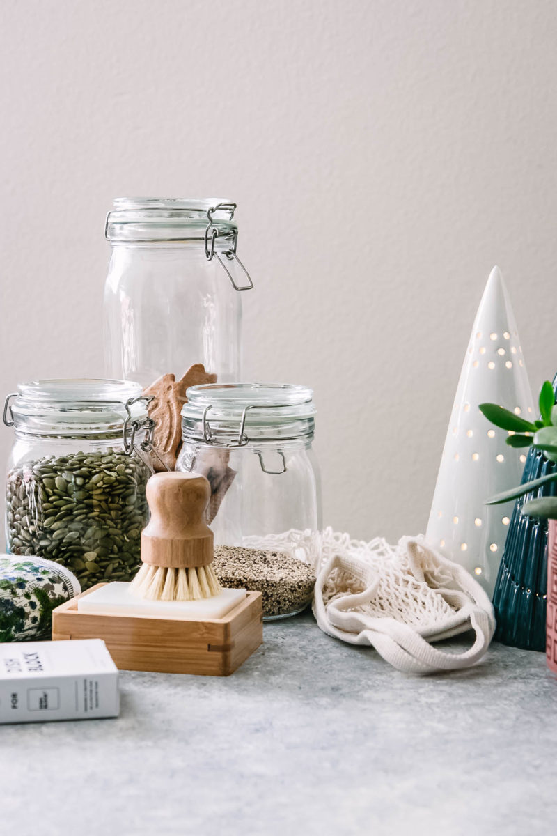 a reusable dish brush, glass food storage jars, and a reusable shopping bag on a table with holiday tree decorations
