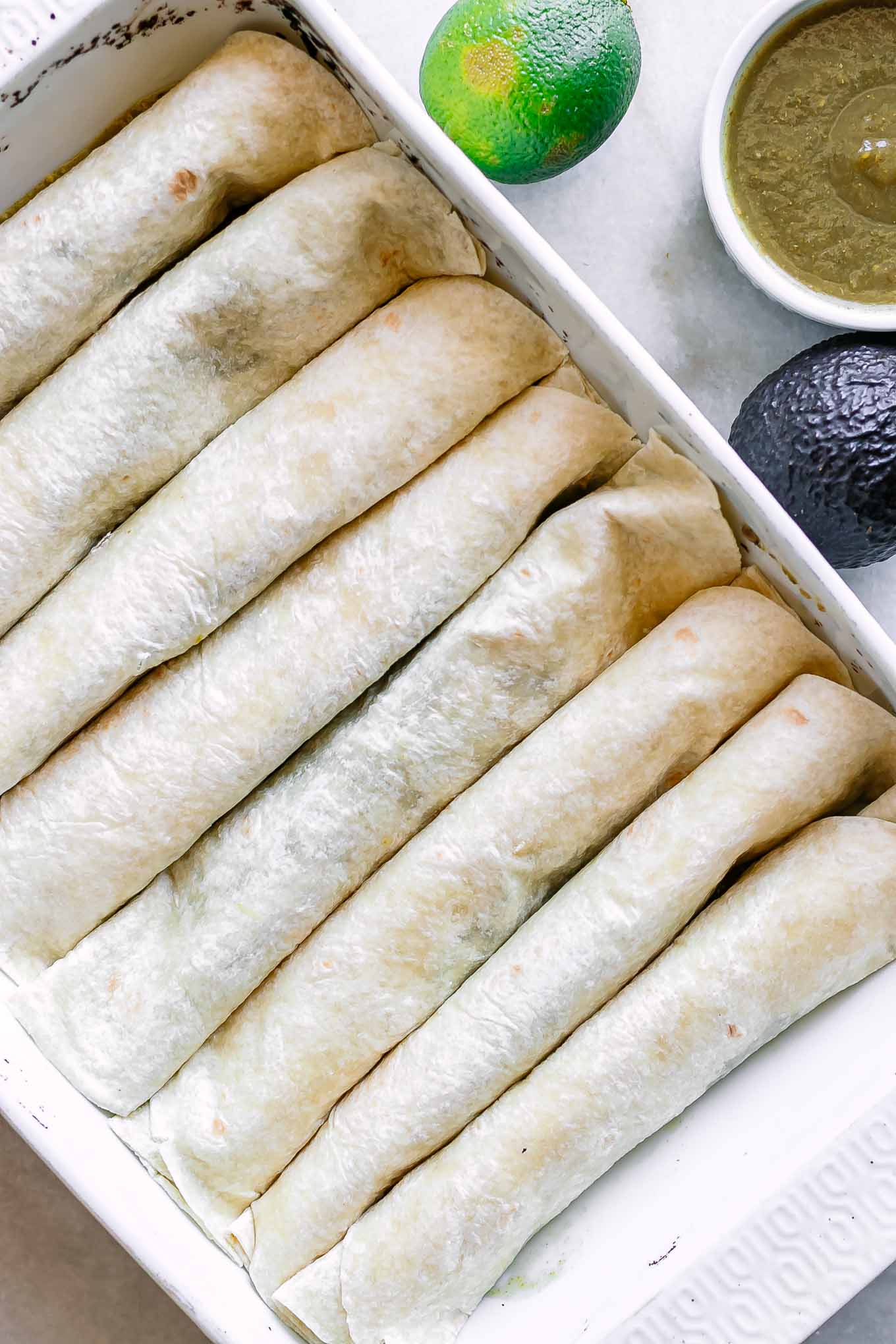 enchiladas in a white ceramic dish on a white table with an avocado and limes