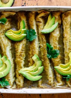 zucchini enchiladas with green enchilada sauce in a white baking dish and garnished with avocado slices and cilantro
